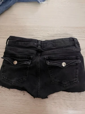 Svarta jeansshorts med rå kant - Snygga svarta jeansshorts med råa, fransiga kanter och silverfärgade knappar. Shortsen har klassiska fem fickor och bakfickor med lock och knapp. Perfekta för en avslappnad och trendig stil.