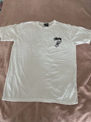 Vit Stüssy dice T-shirt  - Vit t-shirt från Stüssy med svart logga och coolt tärningsmotiv på bröstet. Klassisk passform och rund halsringning, perfekt för en streetwear-look. Materialet känns mjukt och skönt mot huden.