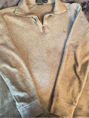 Polo Ralph lauren quarter zip grå - Snygg grå långärmad tröja från Polo Ralph Lauren med halv dragkedja och klassisk broderad logga på bröstet. Tröjan har ribbade muddar och krage, perfekt för en stilren och avslappnad look.