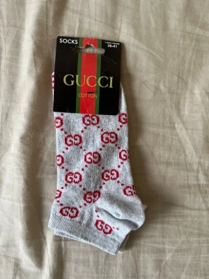 Gråa Gucci strumpor med rött mönster - Snygga gråa strumpor från Gucci med rött GG-mönster över hela strumpan. Tillverkade i mjuk bomull som känns skön mot huden. Perfekta för dig som vill ha en lyxig och trendig detalj i din outfit.
