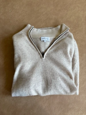 Beige halvzip cashmere tröja från Davida - Mjuk och stilren beige stickad tröja från Davida med halvzip och hög krage. Tröjan har långa ärmar och ribbade muddar vid ärmslut och nederkant. Perfekt för lager-på-lager och en clean look. Nypris ca 2999kr