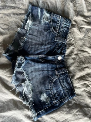 Lågmidjade jeansshorts från Ginatricot - Snygga mörkblå jeansshorts med råa, fransiga kanter och slitna detaljer framtill. Klassisk femficksmodell med knapp och dragkedja. Perfekta för en avslappnad och trendig look.
