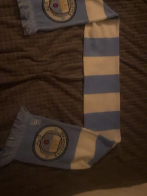 Manchester City halsduk blåvit - Säljer en klassisk halsduk med Manchester Citys klubbmärke. Halsduken har breda blå och vita ränder samt fransar i ändarna. Perfekt accessoar för alla City-fans som vill visa sitt stöd på matchen eller i vardagen.