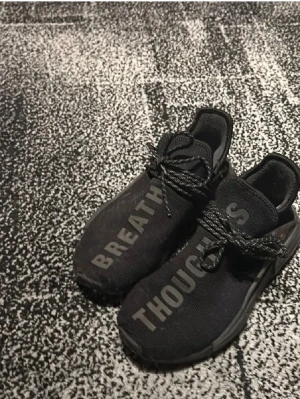 Svarta Pharrell x Adidas - Unika svarta sneakers från Pharrell x Adidas med texten 'BREATHE' och 'THOUGHTS' på ovandelen. Skorna har snörning, stickad ovandel och en chunky sula. Perfekta för dig som gillar streetwear och vill sticka ut. Använda 2 gånger och har där med ett välldigt bra skick 10/10. Boxen skickas med vid köp. Välldigt bra pris då dem ligger på runt 2000kr på andrahands marknaden. 