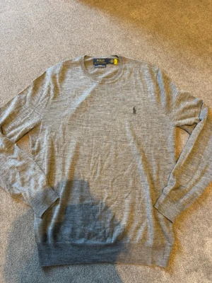 Grå stickad tröja från Polo Ralph Lauren - Snygg grå långärmad stickad tröja från Polo Ralph Lauren med rund halsringning och diskret broderad logga på bröstet. mjukt och tunt material. Välklädd plagg. (Pris kan diskuteras). slim fitt.