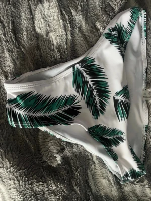 Vit bikiniunderdel med palmblad - Snygg bikiniunderdel i vitt med grönt och svart palmbladsmönster. Klassisk modell med medelhög midja och bekväm passform. Perfekt för strandhäng och pooldagar. Materialet känns mjukt och stretchigt.