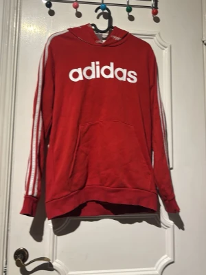 Röd Adidas hoodie med vita ränder - Röd hoodie från Adidas med stor vit logga på bröstet och klassiska vita ränder längs ärmarna. Utrustad med huva och känguruficka framtill. Perfekt för en sportig och avslappnad stil.