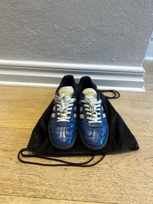 Adidas Samba Wales Bonner Croc - Exklusiva och svårhittade Adidas Samba Wales Bonner Croc i en stilren blå färg med lyxig croc-präglad yta. Modellen kombinerar den klassiska Samba-silhuetten med Wales Bonners unika design och premiumdetaljer. Vita signaturränder och skosnören ger en snygg kontrast mot den mörka sulan. Perfekta för dig som vill ha ett par sneakers som verkligen sticker ut och samtidigt håller hög modegrad.