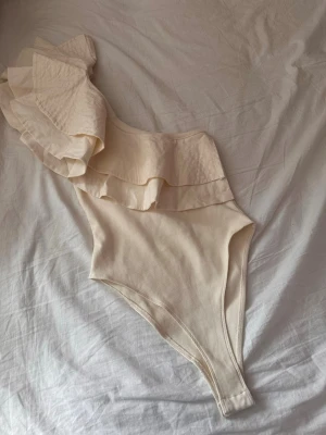  Zara body - Snygg krämfärgad/vit body från Zara med asymmetrisk axel och dubbla volanger. Tillverkad i ett mjukt tyg som känns bekvämt mot huden, i strl M men passar mig som har xs/s. Helt ny, bara att jag tagit av lappen då jag trodde att den skulle komma till användning. Men den ligger bara i min garderob 