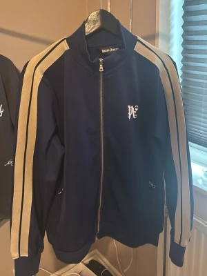 Palm Angels blå track jacket - Snygg blå track jacket från Palm Angels med dragkedja framtill och vita ränder längs ärmarna. Tröjan har hög krage, ribbade muddar och logga på bröstet. Perfekt för en sportig och trendig look.