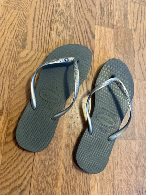 Grå flip-flops från Havaianas - Snygga grå flip-flops från Havaianas med smala remmar i silverfärg och en liten dekorativ detalj på remmen. Klassisk design med mönstrad sula i gummi som ger bra grepp. Perfekta för sommaren eller stranden.