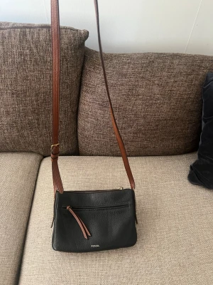 Svart axelväska från Fossil - Stilren svart axelväska från Fossil i slätt läder med justerbar brun axelrem. Väskan har ett ytterfack med dragkedja och dekorativ läderdetalj. Perfekt storlek för det viktigaste och enkel att matcha till olika outfits.