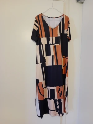Mönstrad långklänning i mjuk polyester - Snygg långklänning med grafiskt mönster i svart, beige och orange toner. Klänningen har korta ärmar, rund halsringning och är tillverkad i ett mjukt polyestertyg. Perfekt för dig som gillar unika prints och bekväma plagg.
