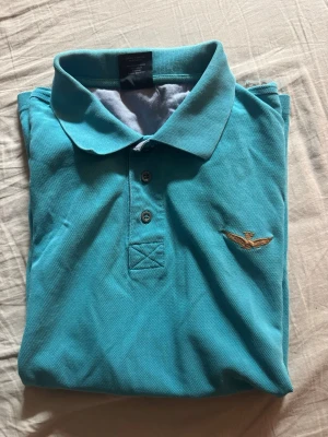 Turkos pikétopp med broderad detalj - Turkos pikétopp med klassisk krage och knappar framtill. Broderad logga på bröstet och rund broderad detalj vid nederkanten. Perfekt för en avslappnad och sportig look.