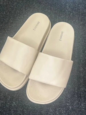 Tofflor HM - Säljer ett par stilrena beigea tofflor från HM. De har en bred rem över foten och är gjorda i ett mjukt, flexibelt gummimaterial. Perfekta för att glida runt hemma eller på stranden. Enkel och modern design som passar till det mesta. Använda 2gånger.