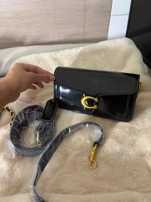 Svart lackad handväska från Coach - Snygg svart handväska i lackat läder från Coach med guldiga metalldetaljer och ett stort C-spänne framtill. Väskan har avtagbar axelrem och kortare handtag, perfekt för att bära på olika sätt. Klassisk rektangulär form och stilren design.