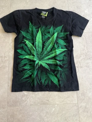 Svart weed tröja - Cool svart t-shirt från The Mountain med stort grönt bladtryck på framsidan. T-shirten har rund hals och korta ärmar. Perfekt för dig som gillar unika och iögonfallande motiv. Materialet känns mjukt och bekvämt mot huden. Jätte bra om du gillar weed