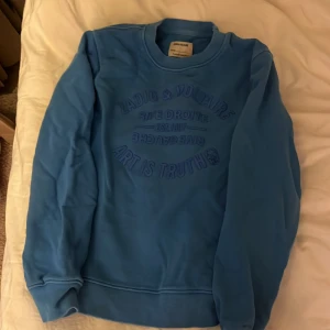 Blå sweatshirt från Zadig & Voltaire - Säljer en blå sweatshirt från Zadig & Voltaire med präglad text på bröstet. Tröjan har rund hals, långa ärmar och ribbade muddar. Perfekt för dig som gillar streetstyle och vill ha något snyggt och bekvämt med coolt tryck.