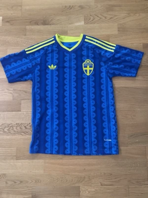 Blå Sverige fotbollströja Adidas - Säljer en blå fotbollströja från Adidas med Sveriges landslagsemblem på bröstet och gula detaljer. Tröjan har mönstrade ränder, gula axelband och texten 'Sverige' i nacken. Perfekt för dig som vill stötta landslaget med stil.