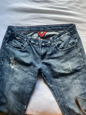 Blå slitna jeans från Y&M - Snygga blå jeans från Y&M med slitna detaljer och klassisk femficksmodell. Jeansen har en cool badge bak med texten 'THE LOVE BRAND' och dekorativa nitar. Perfekt för dig som gillar en avslappnad och trendig stil.