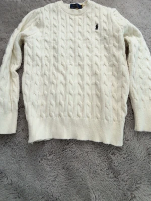 Vit kabelstickad tröja Polo Ralph Lauren - Kabelstickad vit tröja från Polo Ralph Lauren med rund halsringning och broderad logga på bröstet. Tröjan har ribbstickade muddar vid ärmslut och nederkant, vilket ger en klassisk och stilren look. Perfekt för lager-på-lager och enkel att matcha.