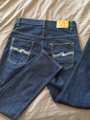Helt nya Nudie Jeans - Säljer ett par klassiska blå jeans från Nudie Jeans med raka ben och snygga kontrastsömmar. Bakfickorna har den ikoniska vita vågsömmen och gult broderi. Tillverkade i slitstark denim, perfekta för en avslappnad och trendig look.
