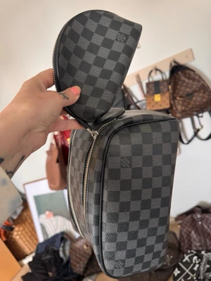 necessär  - Snyggt necessärset i klassiskt grått och svart damier-mönster. Setet består av en större necessär och en mindre rund väska, båda med silverfärgade dragkedjor. Tillverkade i tåligt canvasmaterial med lyxig känsla.