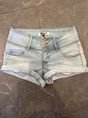 Ljusblå denimshorts från ONLY - Säljer ett par ljusblå jeansshorts från ONLY med låg midja och och kort ben. Shortsen har tre knappar framtill och snygga sömdetaljer. Perfekta för sommaren. De är nyskick och aldrig använda❣️