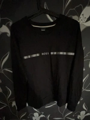 Svart sweatshirt från BOSS - Cool svart sweatshirt från BOSS med silverfärgad logga och dekorativ rand över bröstet. Tröjan har rund halsringning och långa ärmar, perfekt för en avslappnad och stilren look. Materialet är mjukt och bekvämt, idealiskt för vardagsbruk.
