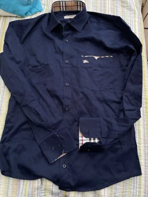 Mörkblå skjorta från Burberry - Snygg mörkblå skjorta från Burberry med klassiskt rutigt tyg på insidan av kragen och manschetterna. Skjortan har lång ärm, bröstficka med broderad logga och knappar framtill. Perfekt för dig som gillar stilrena detaljer och exklusiva märken.