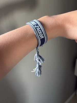 Blått J'Adior armband från Dior - Snyggt blått tygarmband från Dior med texten 'J'Adior' invävd i vitt. Armbandet har justerbar knytning med tofsar i ändarna och en cool, bohemisk känsla. Perfekt accessoar för att lyfta din outfit med en touch av lyx.