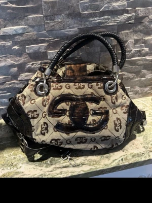 Beige och brun handväska - Snygg handväska i beige canvas med brunt -mönster och stora svarta detaljer framtill. Väskan har svarta handtag i flätad look och silverfärgade metalldetaljer. Perfekt för dig som gillar statement-accessoarer.
