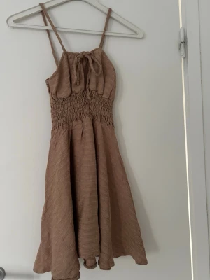 Beige sommarklänning med smock - Söt beige klänning med smala axelband och smock i midjan. Klänningen har en rosett framtill och ett lätt, luftigt tyg som passar perfekt för varma dagar. 