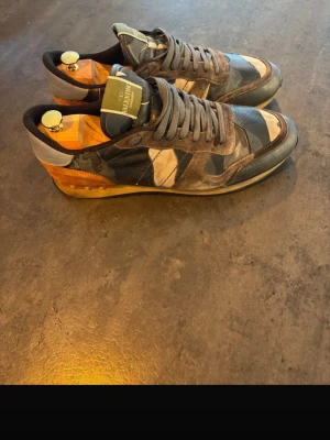 Valentino rockrunners  - Unika sneakers från Valentino med camo-mönster i blått, grått och svart. Skorna har detaljer i mocka och läder samt en cool orange häl. Klassisk låg modell med snörning och robust sula. Perfekta för dig som vill sticka ut med din stil.