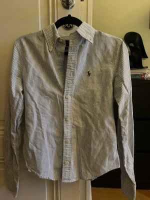 Randig skjorta från Ralph Lauren - Klassisk skjorta från Ralph Lauren med smala blå och vita ränder. Skjortan har lång ärm, knappar framtill och button-down krage. Broderad logga på bröstet. Perfekt för en stilren och tidlös look.