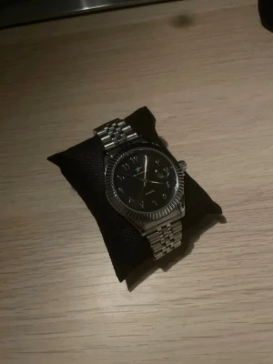 Silverfärgad klocka med arabiska siffror  - Stilren klocka med silverfärgad länk i metall och svart urtavla. Klockan har tydliga index och datumvisning, samt klassisk rund form. Perfekt accessoar för dig som gillar en clean och tidlös look.