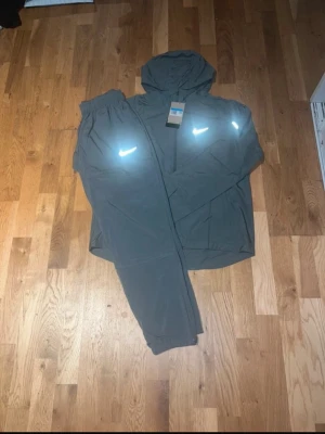Nike grå träningsset med huva - Säljer ett grått träningsset från Nike med matchande byxor och jacka med huva. Setet har reflekterande Nike-loggor på både jackan och byxorna. Perfekt för träning utomhus och har en sportig, avslappnad passform.