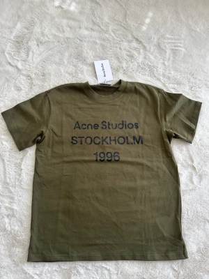Olivgrön Acne Studios t-shirt - Säljer en olivgrön t-shirt från Acne Studios med tryck i svart text på bröstet: 'Acne Studios STOCKHOLM 1996'. Klassisk passform med rund hals och korta ärmar. Tillverkad i mjuk bomull, perfekt för en clean och avslappnad stil.