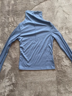 Ljusblå polotopp med långa ärmar - Snygg ljusblå polotopp med lång ärm och hög krage. Perfekt att styla med jeans eller kjol för en clean look. Mjuk och stretchig känsla i materialet.
