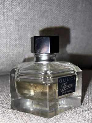 Gucci Flora Eau de Toilette parfym - Gucci Flora Eau de Toilette i en elegant, sexkantig glasflaska med svart etikett och svart lock. Parfymen har en ljusgul vätska och flaskan känns lyxig och stilren – perfekt för dig som gillar exklusiva dofter.