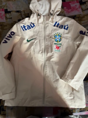 Vit Brasil windbreaker  - Vit vindjacka från Nike med huva, officiell Brasilien landslagsmodell. Jackan har tryckta loggor från sponsorer som Itaú och Vivo samt Brasilien-emblem på bröstet. Dragkedja framtill och justerbar huva. Perfekt för fotbollsfans som vill sticka ut.