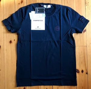 Mörkblå t-shirt från Stone Island - Snygg mörkblå t-shirt från Stone Island med klassisk rund hals och korta ärmar. Diskret logga broderad på bröstet och en liten detalj med knapp på ärmen. Perfekt för en stilren och avslappnad look.