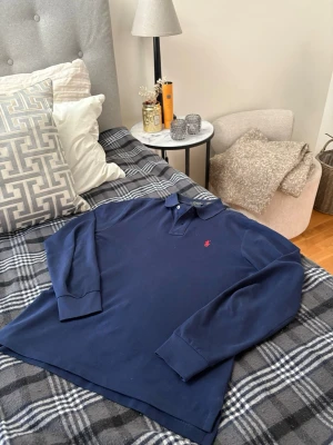 Polo Ralph Lauren långärmad piké - Klassisk marinblå långärmad pikétröja från Polo Ralph Lauren med röd broderad logga på bröstet. Skjortan har krage, knappar vid halsen och är tillverkad i mjuk bomull. Perfekt för en stilren och avslappnad look.
