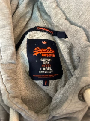 Grå hoodie från Superdry med tryck - Snygg grå hoodie från Superdry med stort mörkblått tryck på bröstet och siffran 54 på magen. Klassisk känguruficka och vita dragsnören i huvan. Mjuk bomullsblandning som är skön att ha på sig. Storlek XL sitter mer som L enligt mig.