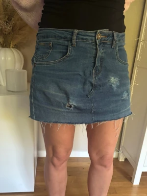 Blå jeanskjol med fransig kant - Trendig blå jeanskjol med fransig, rå kant och slitna detaljer framtill. Klassisk femficksmodell med knapp och dragkedja. Perfekt för en avslappnad och cool stil.