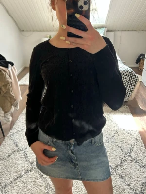Svart basic kofta med knappar - En svart, enkel kofta med rund halsringning och knäppning framtill. Långärmad modell i mjukt material som passar perfekt till jeans eller kjol. Stilren och lätt att matcha för en clean look. Blomrig pattern på framsidan, normal på baksidan.