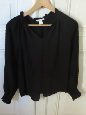 Svart blus med struktur från H&M - Snygg svart blus från H&M med diskret prickig struktur i tyget. Blusen har v-ringning, långa ärmar med smock vid ärmslut och lätt rynkad halsringning🌸