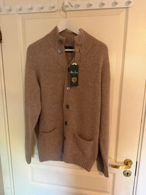 Beige kofta från Alan Paine i lammull - Mysig beige kofta från Alan Paine i 100% lammull. Koftan har knäppning framtill, två fickor och ribbstickade muddar. Perfekt för lager-på-lager och har en klassisk, tidlös look med hög krage och snygga detaljer.