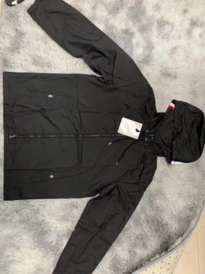 Svart vindjacka från Moncler - Svart vindjacka från Moncler med huva och dragkedja framtill. Jackan har två fickor med tryckknappar och diskreta detaljer i rött, vitt och blått på insidan av huvan. Tillverkad i lätt och smidigt material, perfekt för blåsiga dagar.