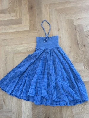 Blå halterneckklänning med smock - Snygg blå klänning med halterneck och smockad överdel. Kjolen är vid och luftig med subtilt mönster. Flätat band i halsen med pärldetaljer. Perfekt för varma dagar och sommarens äventyr.